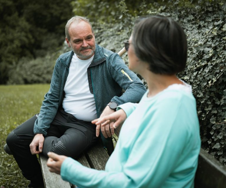 Coachingsession sitzend Mann und Frau sitzen auf einer Bank und unterhalten sich in einer grünen Umgebung.