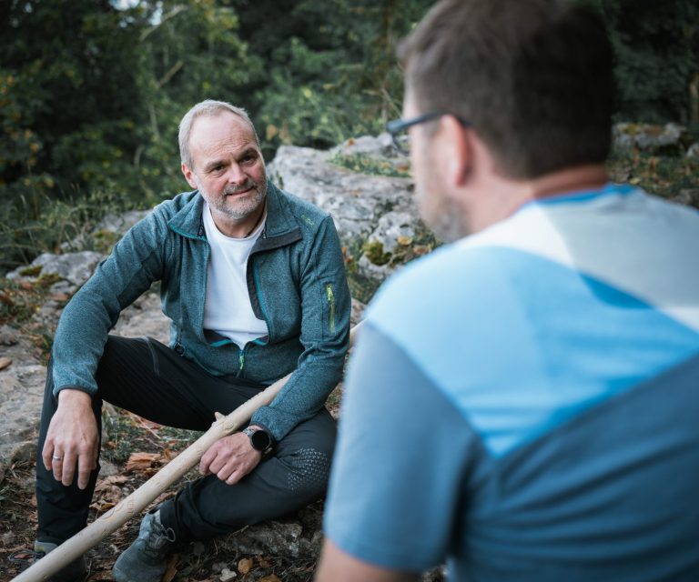 Einzelcoaching Zwei Männer im Gespräch, einer sitzt auf einem Felsen, der andere steht ihm gegenüber.