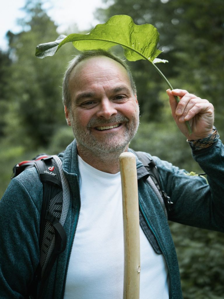 Mann mit einem Blatt als Hut, lächelt in einem Wald. Trägt einen Rucksack.