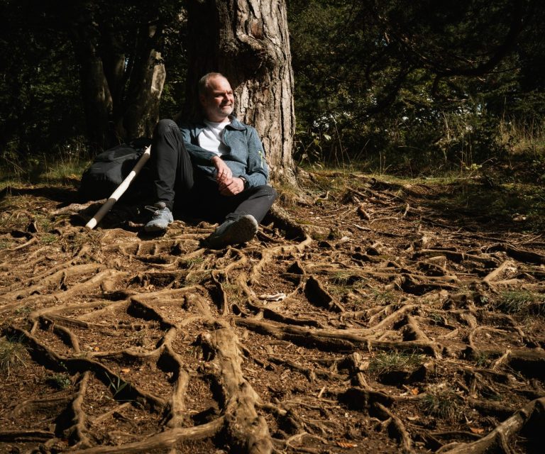 Naturcoach Dominic Zäh auf Wurzeln Mann sitzt entspannt an einem Baum, umgeben von Wurzeln und natürlichem Licht.