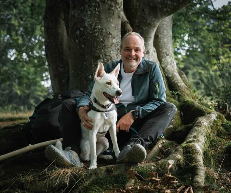 Naturcoach Dominic Zäh mit Zoe Ein Mann sitzt mit einem weißen Hund an einem Baumstamm im Freien.